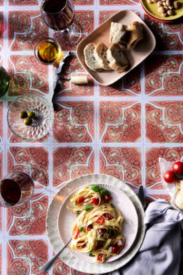 Tablescape with pasta and tomatoes, olives, bread, glasses of wine, and a plate of marcona almonds